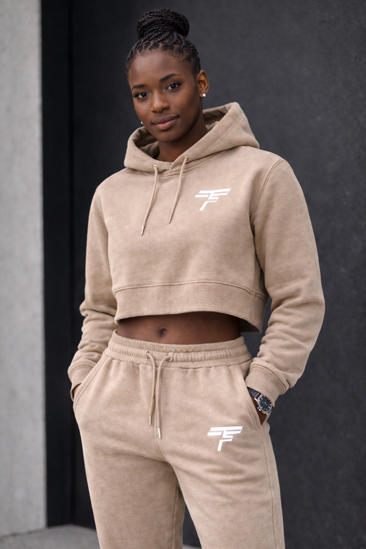 Woman wearing a beige hoodie and pants with a brand logo against a dark background